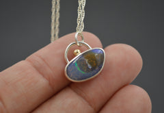 Unique boulder opal pendant that resembles a sandy beach  island with blue water and green line detail. Green gold dot detail on the top. On a silver chain resting on fingertips
