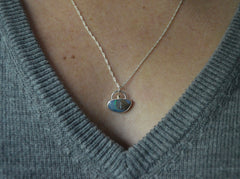 xUnique boulder opal pendant that resembles a sandy beach  island with blue water and green line detail. Green gold dot detail on the top. On a silver chain worn on model with gray sweater.