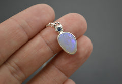 Pendant with an Australian Opal with a blue Montana sapphire set in a star on top of it. On a silver chain laying on top of fingertips  Edit alt text