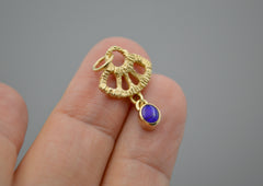 Gold textured fan shape charm with a dark blue opal dangling from the bottom laying on fingers
