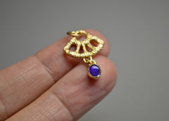 Gold textured fan shape charm with a dark blue opal dangling from the bottom laying on fingers