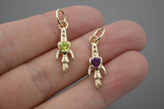 Two gold rocket ship pendants with gemstones held in a hand against a neutral background