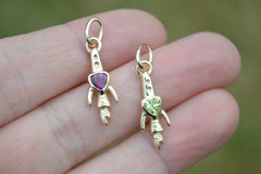 Two small gold charms with gemstones held in a hand against a blurred green background