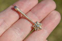 14k yellow gold star wand shaped pendant with a white diamond in the center of the star. Reseting on a hand out in natural sunlight with a green background.