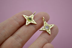 14k yellow gold four pointed star compass pendants. Left one has an olive green tourmaline and the right has a very pale milky blue chalcedony cabochon. Resting on fingers with a pink background.
