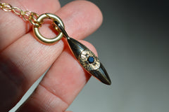 14k gold eye pendant that hangs vertically. The pupil of the eye is a faceted black diamond. Shown on a circular charm holder and chain. Laying on a hand.
