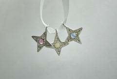three four pointed silver star pendants handing on a ribbon. Left has a berry colored garnet, center has an opal and right has a blue Montana sapphire
