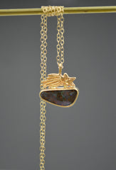 Gold Necklace with a shooting star sitting on top of an Australian boulder opal with the chain hanging and wrapped on a dowel on a gray background