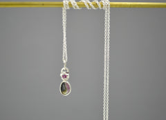 Silver pendant with star element at the top that has a berry pink rhodolite garnet set in the center of it. Underneath is a rose cut bi color tourmaline stone in a bezel setting. The tourmaline has both dark green and baby pink colors in it. Hanging on a chain wrapped around a brass colored bar.