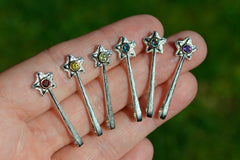 Silver star-shaped wand pendants with colored gemstones held in a hand against a blurred green background